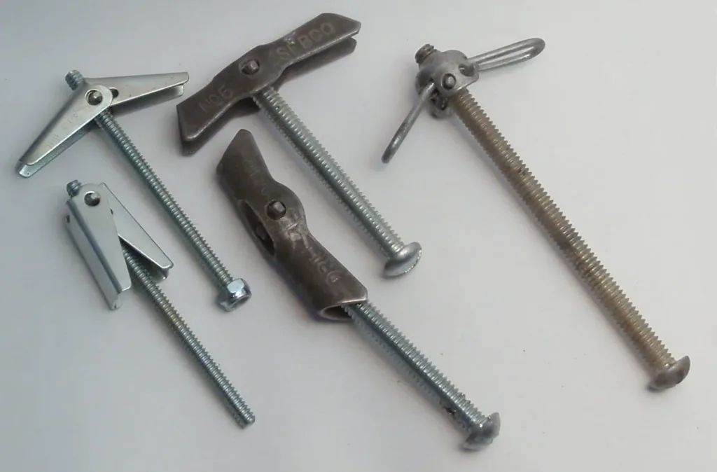 Fixadores metálicos com asas articuladas (tipo toggle) para parede oca, sobre superfície cinza